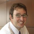 Headshot of a white man with brown hair wearing small rectangular glasses and a white button down shirt and tie. 