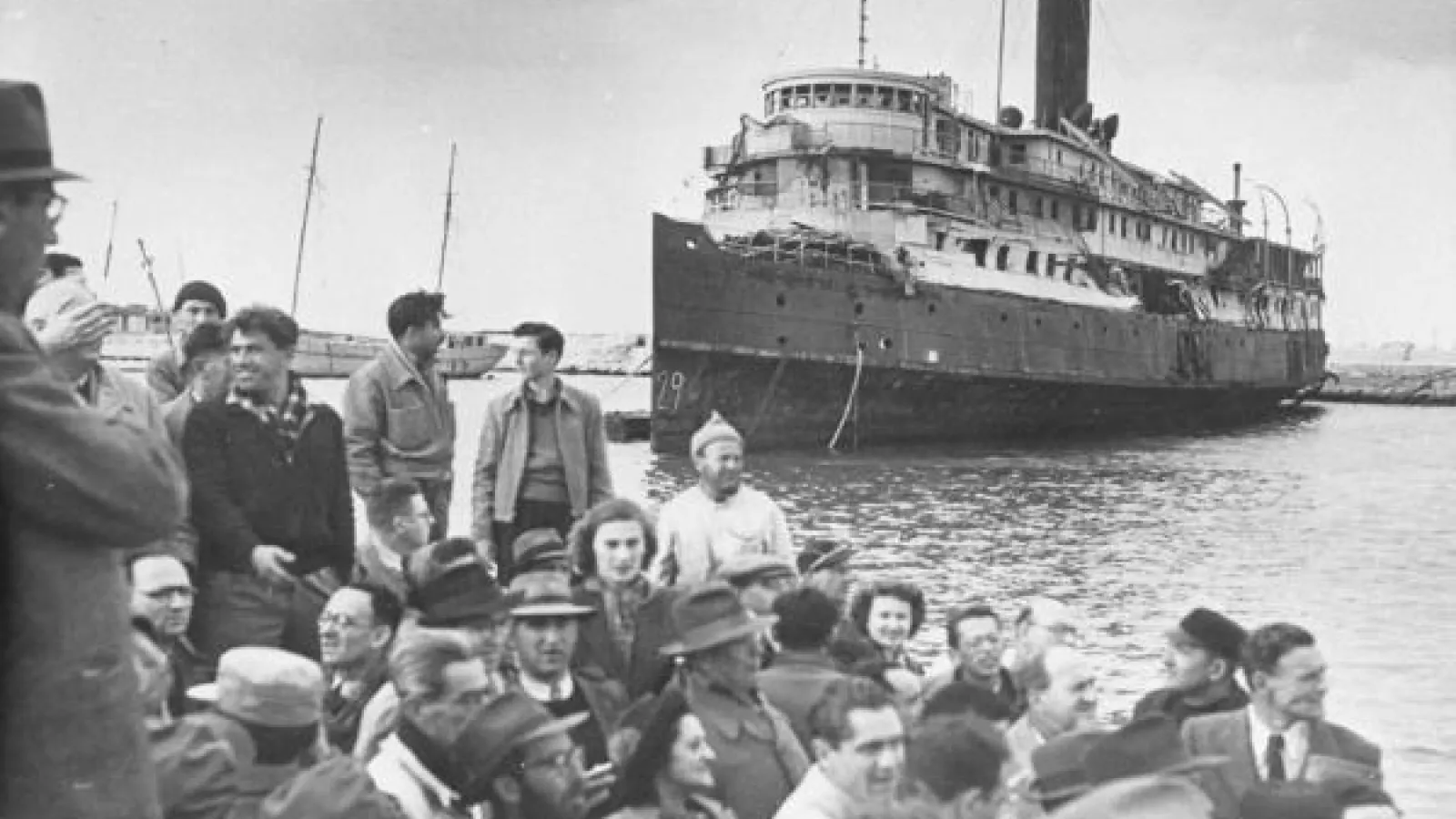 A large ship approaches a group of people standing on a dock