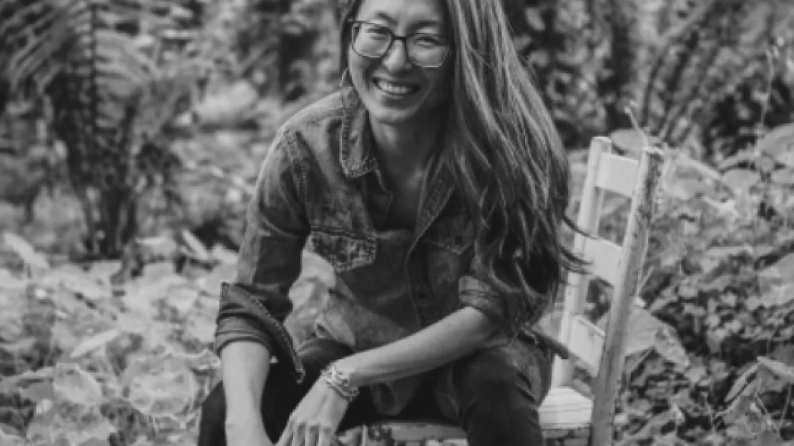 Black and white portrait of Mia Mingus sitting on a wooden chair in a jungle setting. 