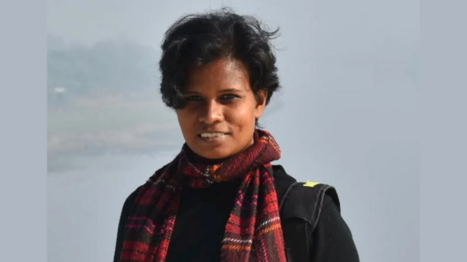 Portrait of the poet with a grey background. She has short brown hair and a plaid red scarf. 