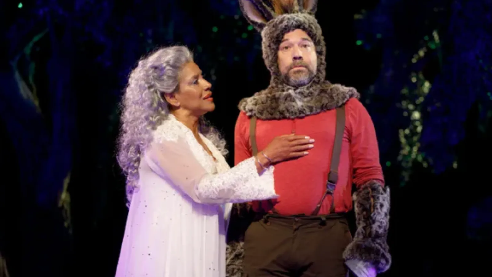 A woman in a long gown touches the chest of a man wearing a furry headpiece with donkey ears