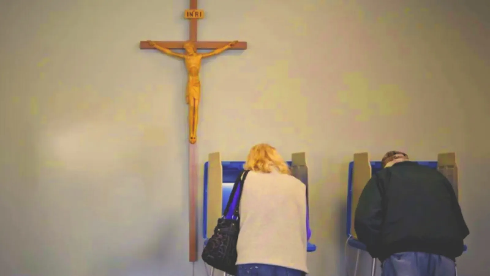 Two people standing a voting booth and a crucifix hangs overhead. 