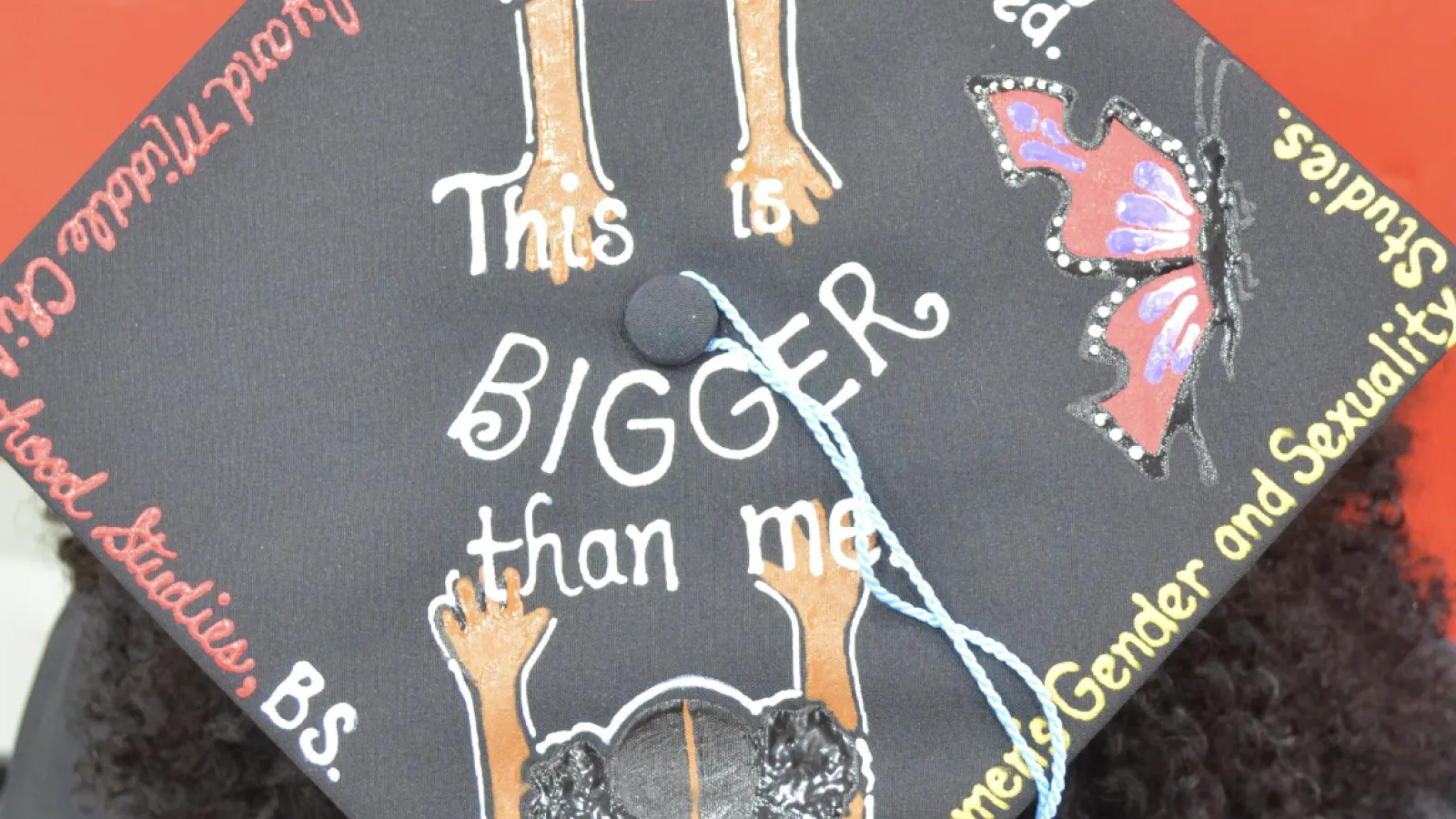 A graduating student wearing a mortarboard cap. In the center, it says "This is BIGGER than me." There are two girls reaching out toward each other from two corners of the bortarboard. The two girls are wearing shirts that say, "Black girl magic" and "I matter." Around the edges of the mortarboar, there is a butterfly and text that reads, "Beautifully flawed," "Women's gender and sexuality studies," "Early and middle childhood studies," "B.S.," and "Kines."