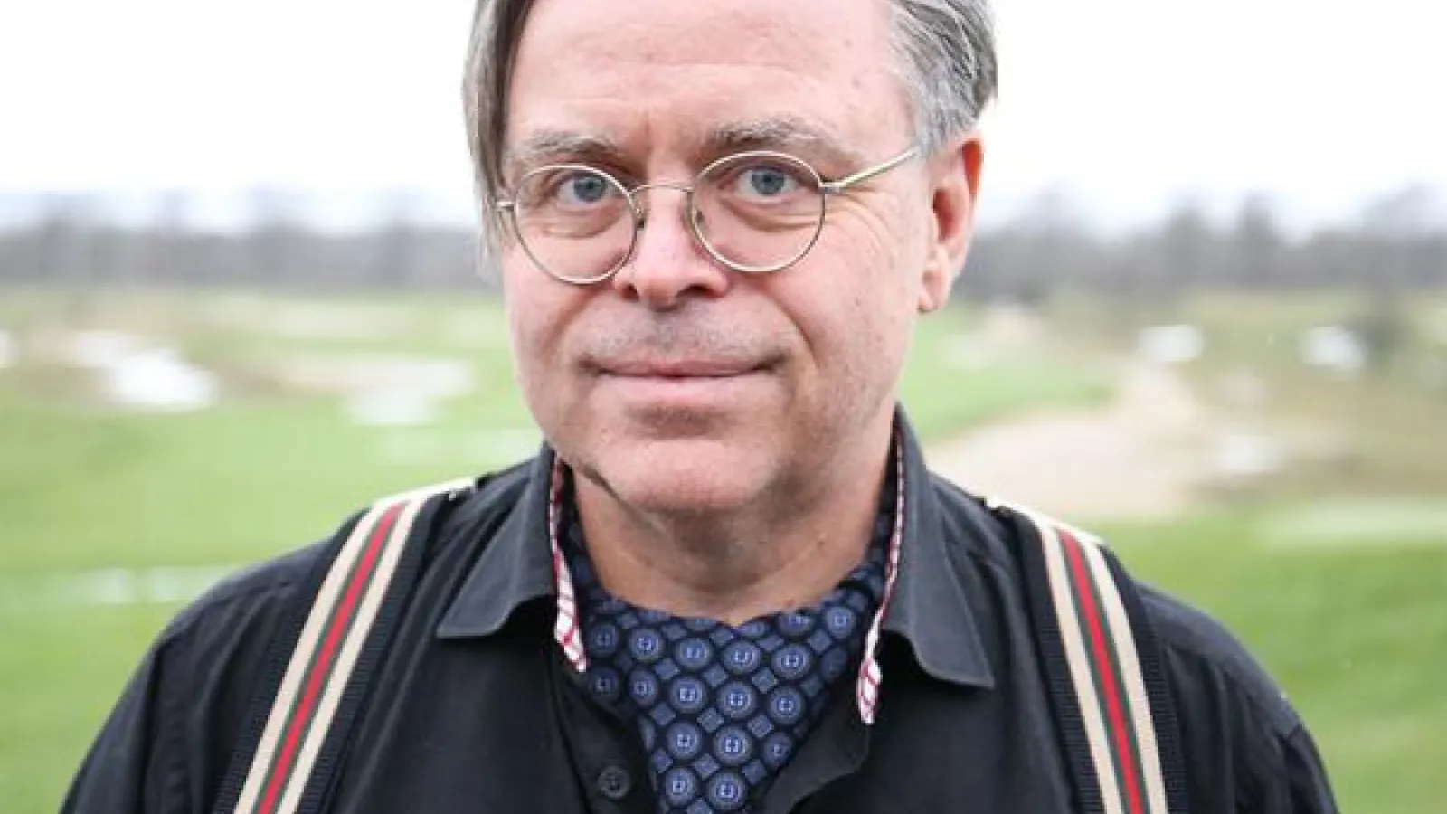 Older man with gray hair, round glasses, wearing a blue button up shirt with striped suspenders