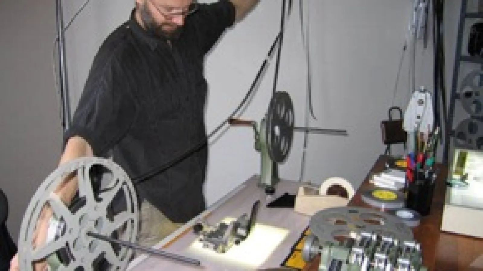 A man standing before a reel to reel projector and he has pulled the film out with widespread arms