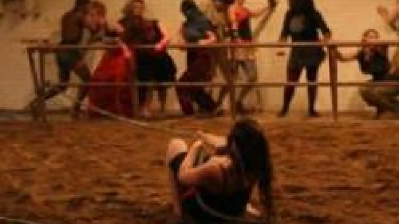 Two women wrestleing in a muddy ring and people are standing around the barrier fence. 