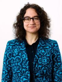 Frontward facing portrait of a woman with black curly hair, black glasses and a blue patterned blazer