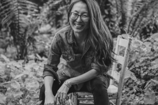 Black and white portrait of Mia Mingus sitting on a wooden chair in a jungle setting.