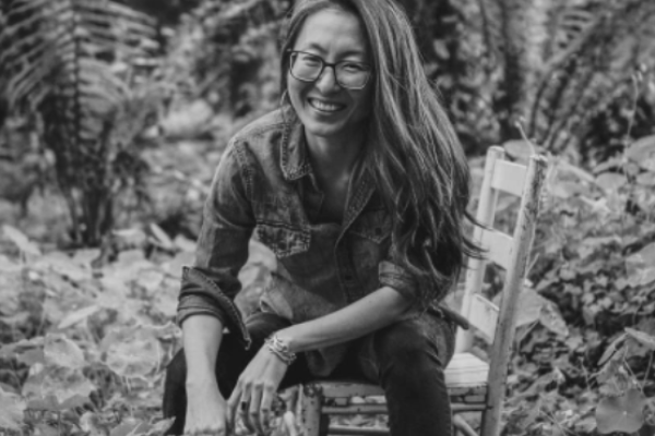 Black and white portrait of Mia Mingus sitting on a wooden chair in a jungle setting. 