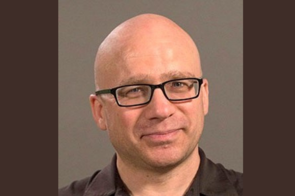 Portrait of a bald white man wearing square black glasses and a slight smile on his face.