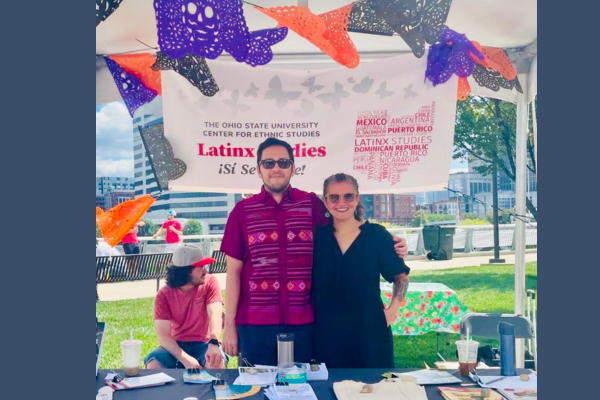 A man and a woman stand at a table, surrounded by papel picado paper flags.