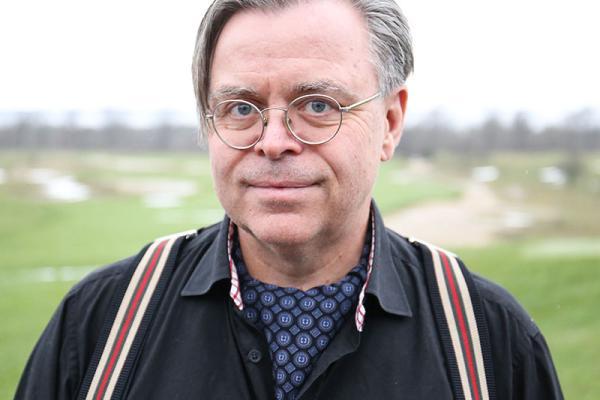 Older man with gray hair, round glasses, wearing a blue button up shirt with striped suspenders