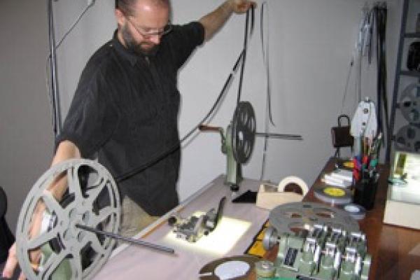 A man standing before a reel to reel projector and he has pulled the film out with widespread arms