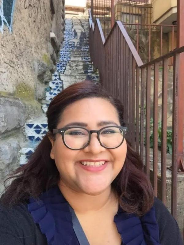 Stephanie Vigil looking at the camera posing in front of a staircase and wearing a black shirt with a blue collar.